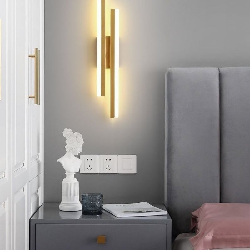 Gold-tone bar wall light above a grey nightstand with a sculpture, books and textured cup