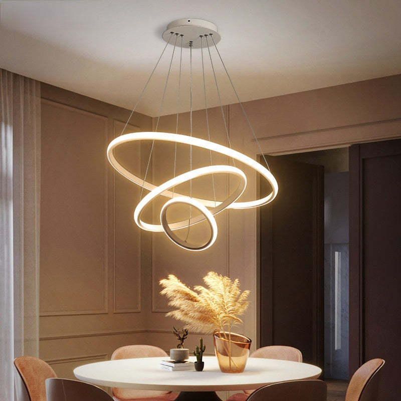 White three-ring pendant light with warm-looking outer illumination above a round dining table and pampas vase