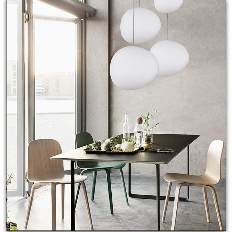 White egg-shaped pendants above a dark dining table with mixed chairs, food and glassware beside a tall window