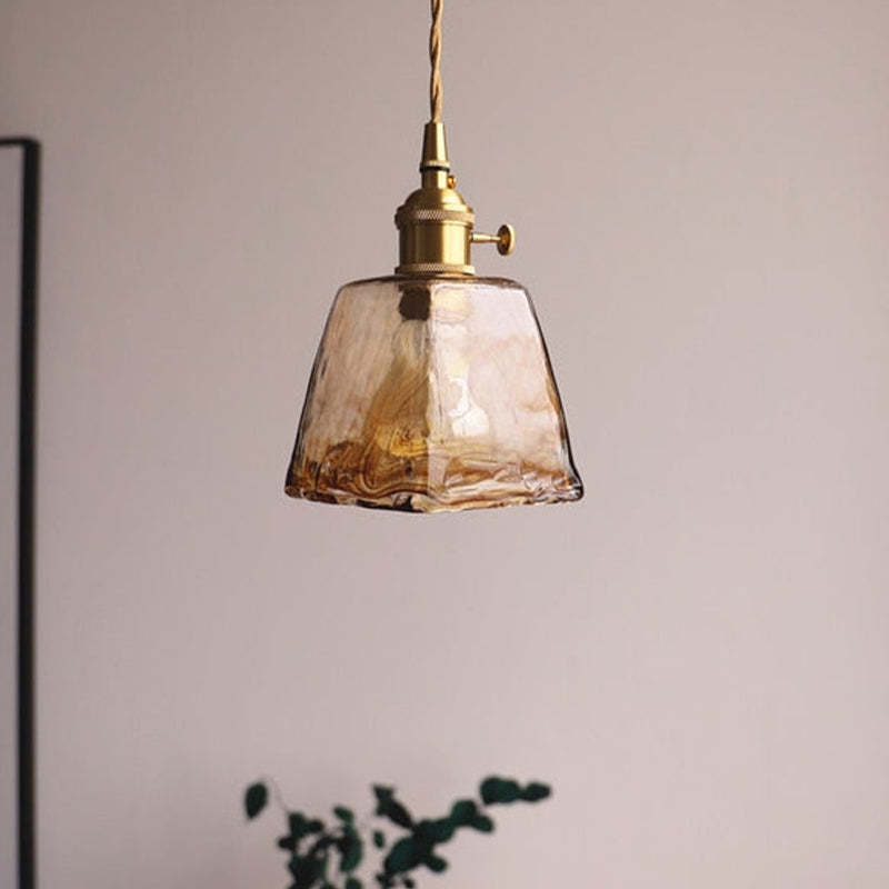 Unlit tapered amber glass pendant with a brass-tone fitting beside a plant