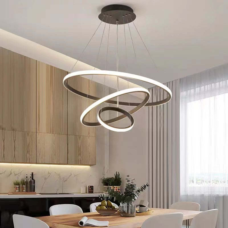 Coffee Brown three-ring pendant light above a wooden table with White chairs, fruit, book and plants beside a kitchen