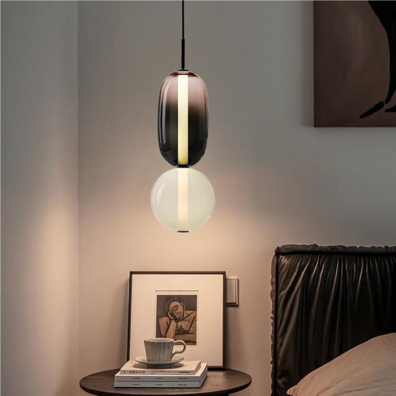 Two-form smoked and pale glass pendant above a bedside table with books, a cup and framed portrait