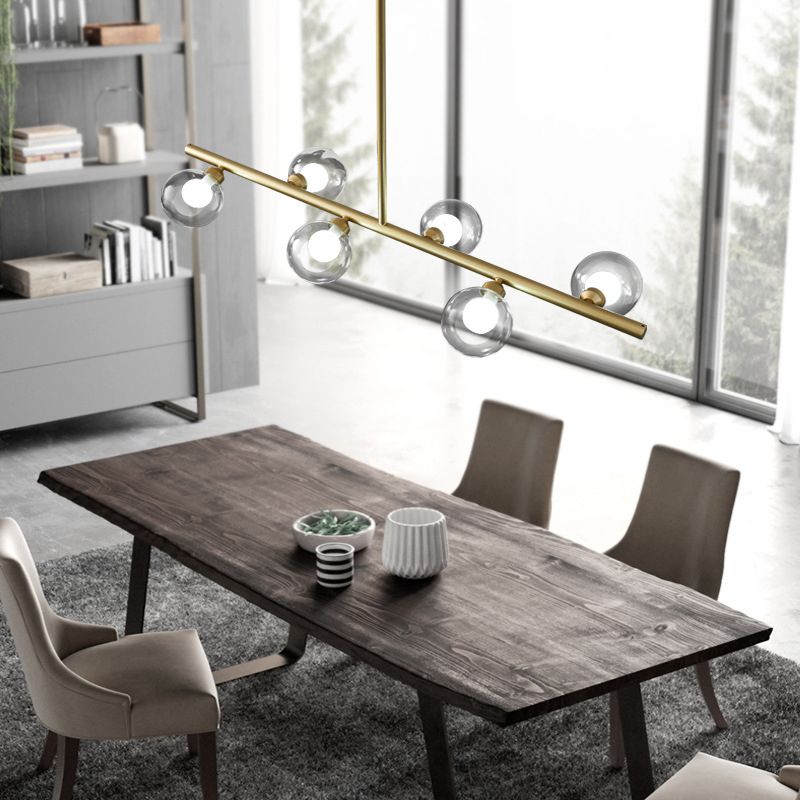 Gold six-globe linear pendant illuminated above a wooden dining table near windows and a bookcase