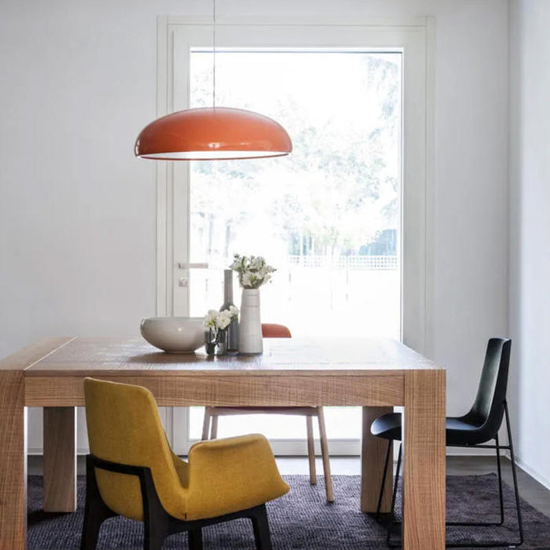 Orange dome pendant over a wooden dining table with vases and a mustard chair beside a window