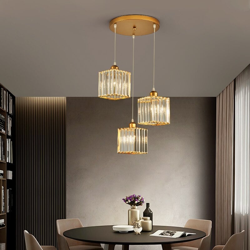 Gold three-square-shade cluster staggered below one round canopy above round dining table, beige chairs and bookcase
