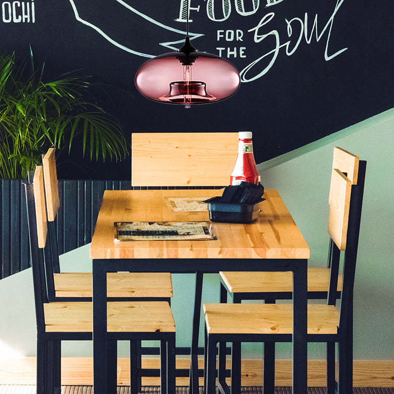 Pink oval glass pendant light above a wooden dining table with Black chairs, menus, napkins and a potted plant