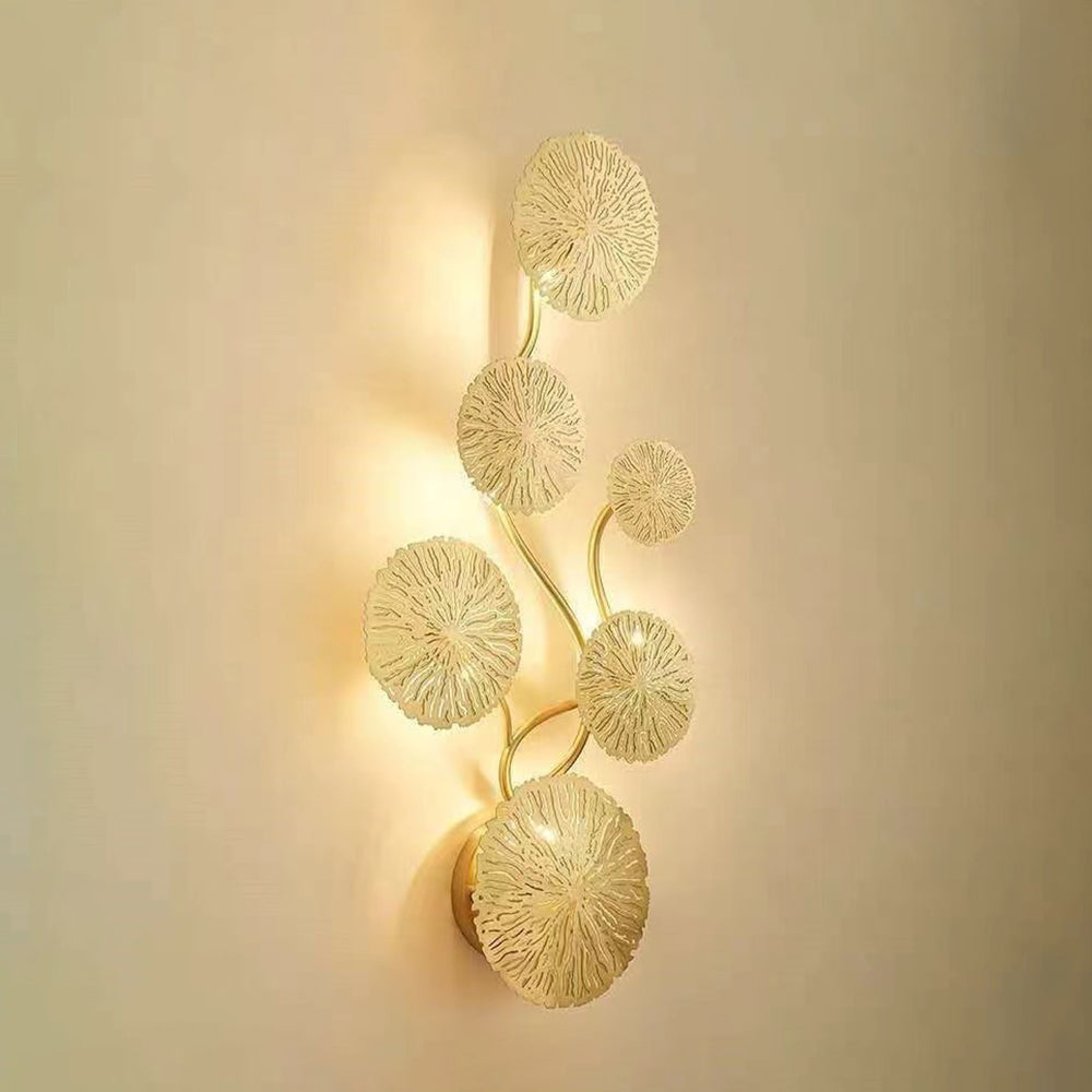 Six-leaf gold-tone wall light with radial texture and a glowing outline on a pale wall