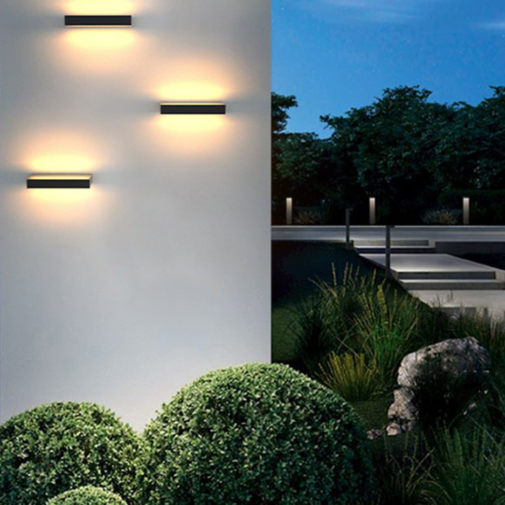 Three dark wall lights arranged horizontally on a pale exterior wall beside a landscaped garden