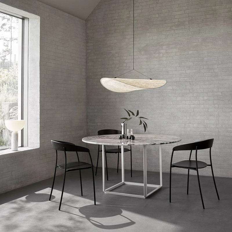 Single white curled pendant over a round stone-look dining table with black chairs beside a large window