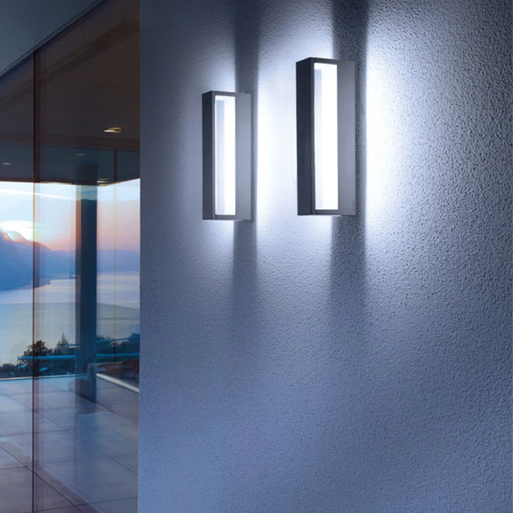 Two upright dark rectangular wall lights with pale illuminated openings beside a glass wall