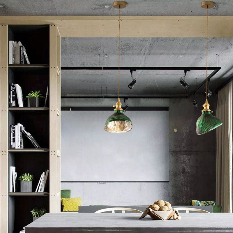 Two green curved-ribbed pendants above a dining table with a bread bowl, beside wood shelving and a concrete ceiling
