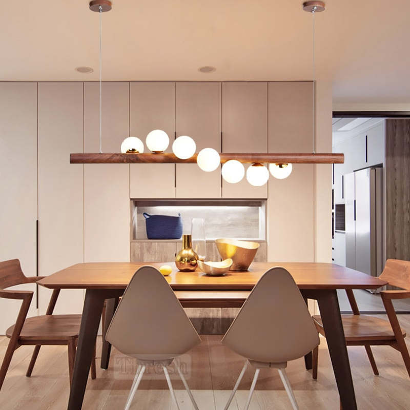 Dark wooden bar pendant with seven White globes above a dining table, Gray chairs, Gold-toned bowls and fitted cabinets