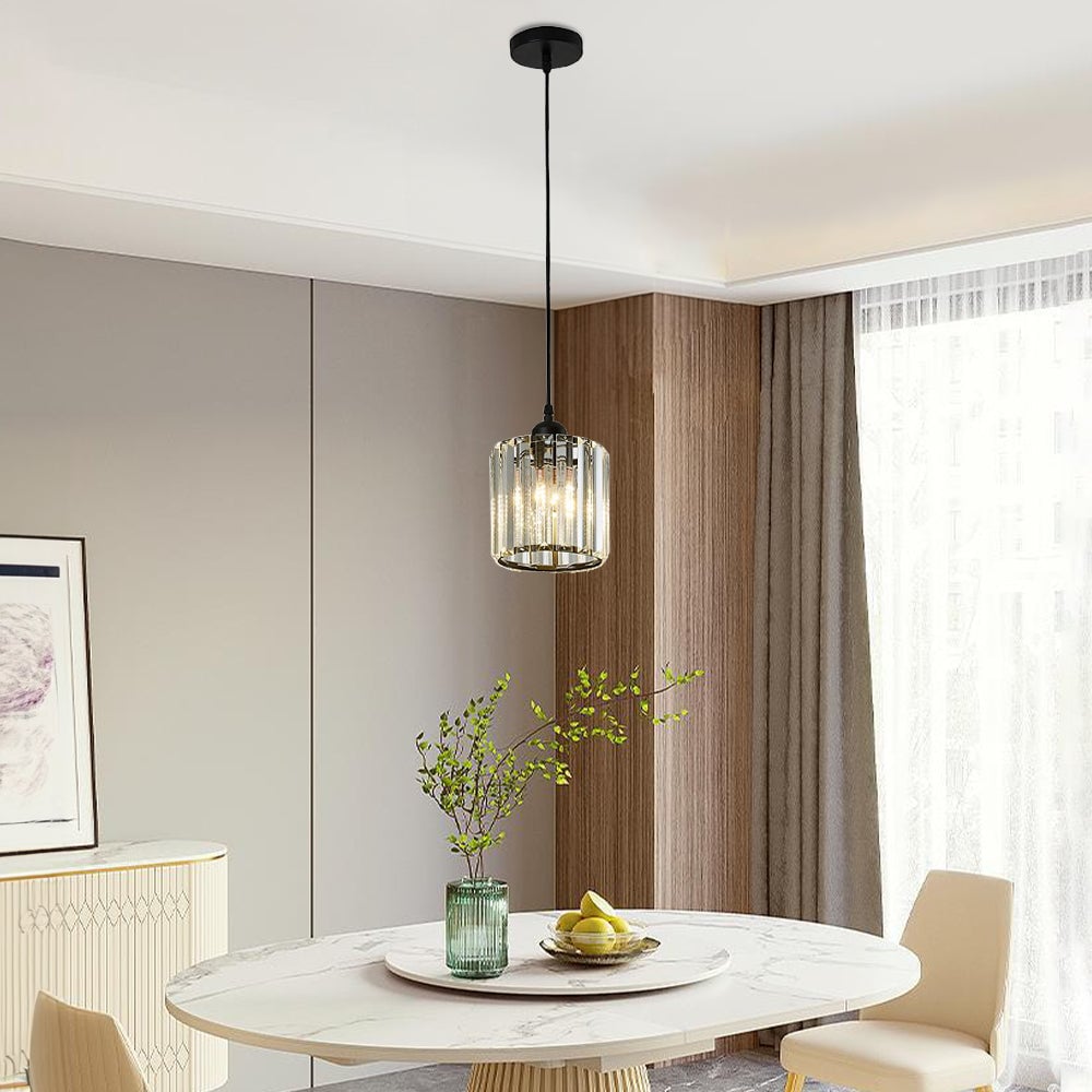 Single black cylindrical glass pendant over round pale table with green vase and fruit beside sheer curtains