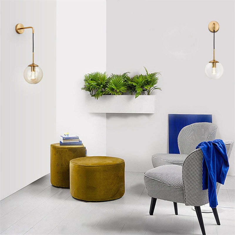 Two gold-finish glass globe wall lights on a seating-area wall with plants and patterned furniture