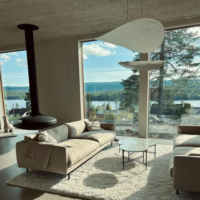 Two pale curled fabric pendants beside landscape windows, cream sofas and a suspended black fireplace