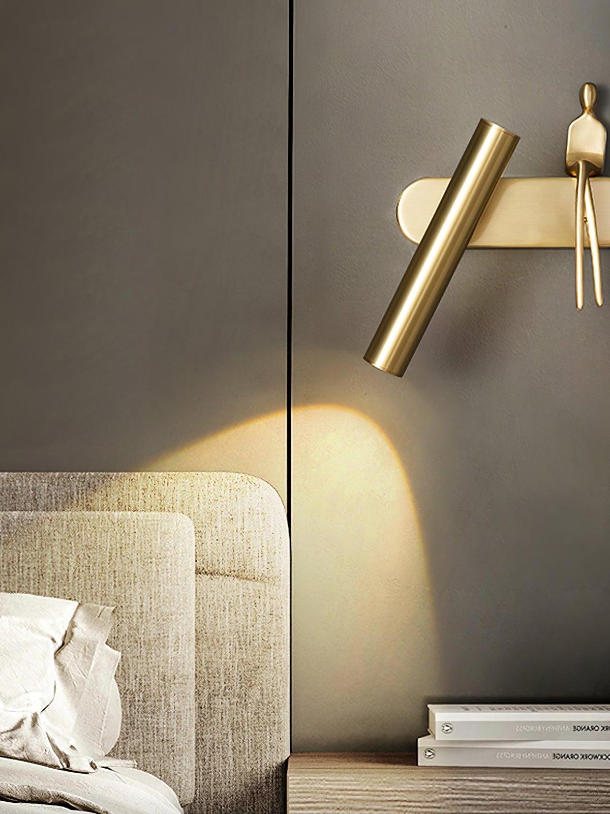 Brass-coloured tube wall light with seated figure above a pale bedhead and wooden bedside table