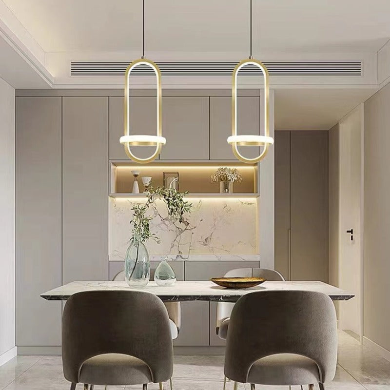 Two gold Nyla oval-and-ring pendants above a rectangular dining table with dark chairs and two green glass vases