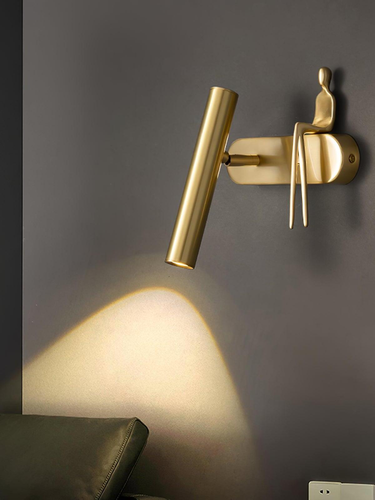 Angled brass-coloured wall light with rounded wallplate, tilted tube and seated figure casting light down the wall