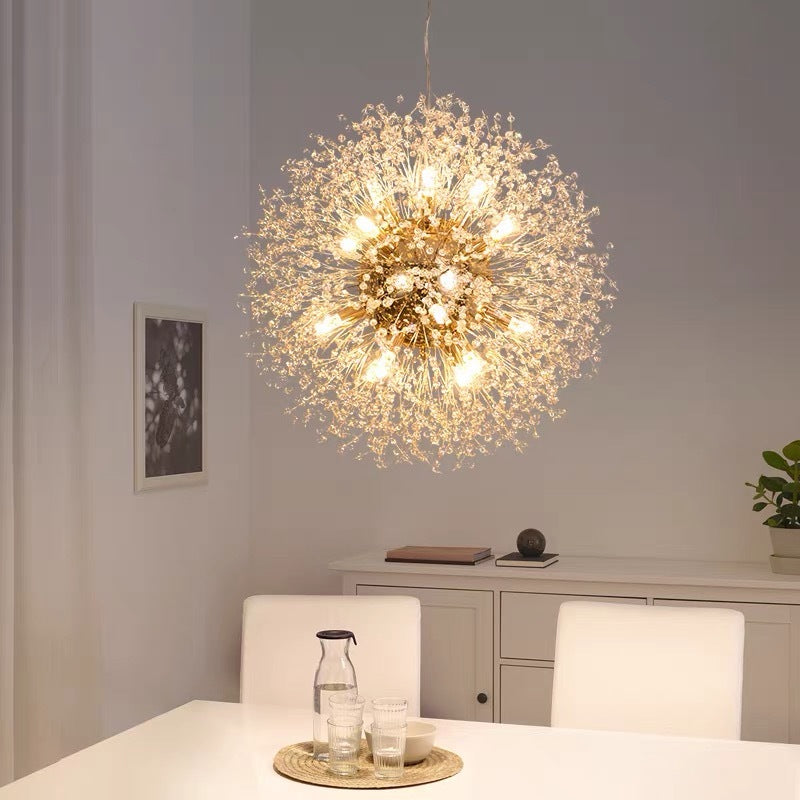 Lit gold Lili dandelion pendant above white dining table with carafe and glasses, white chairs and sideboard with books and art
