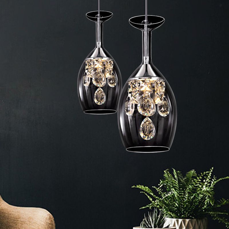 Two elongated clear Kristy pendants with faceted hanging drops beside a chair and potted plants against a dark wall.