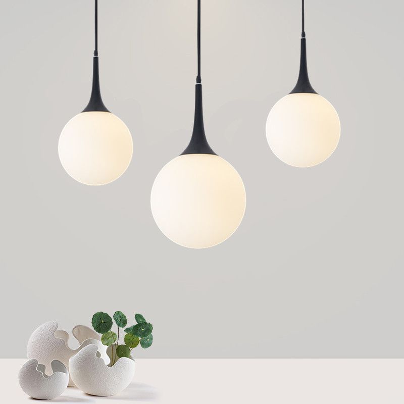 Three glowing white Valentina globes with black tapered necks at staggered heights above sculptural white vases and green leaves