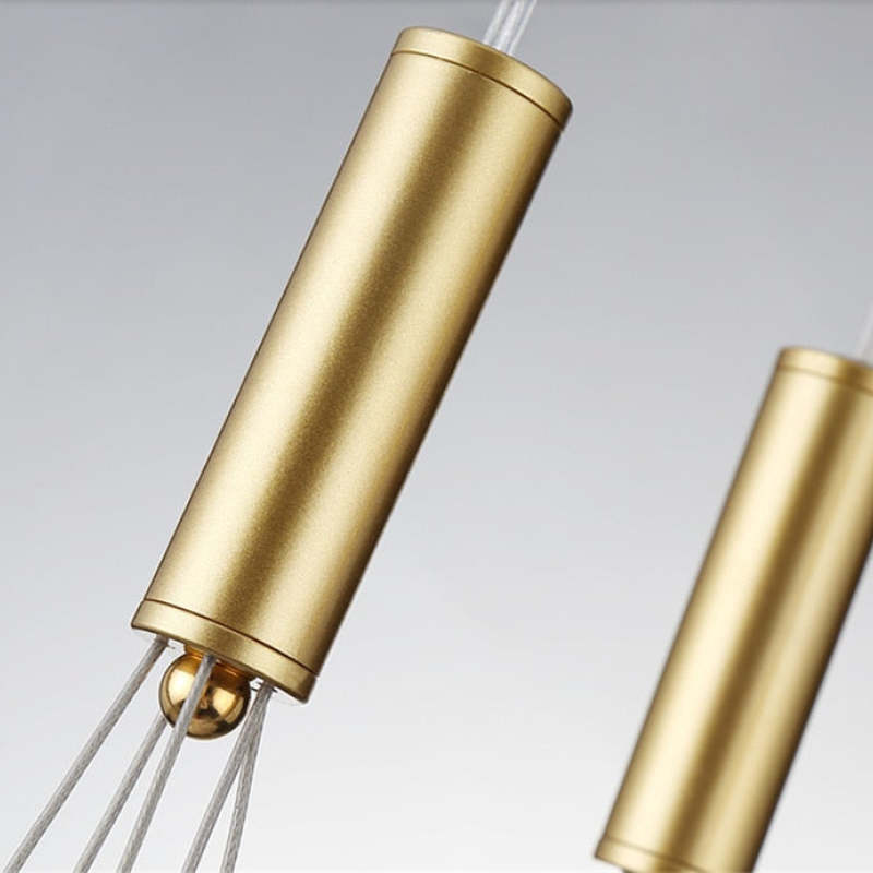 Close-up of a Valentina gold-tone cylindrical fitting, shiny junction and pale hanging wires, with a second fitting blurred