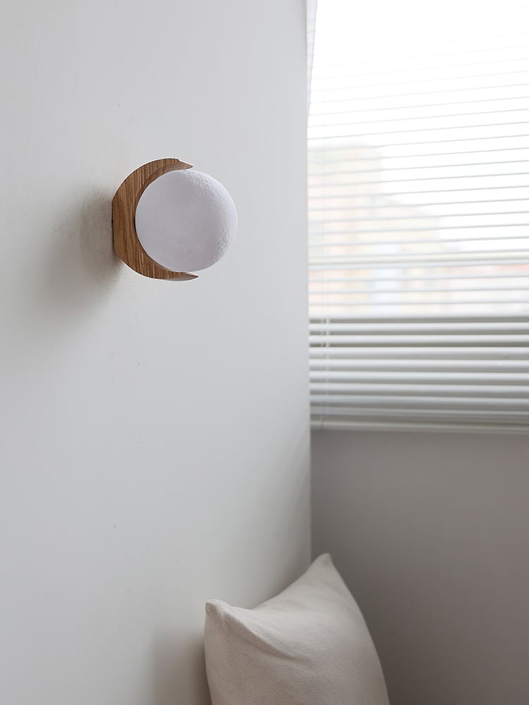 Unlit white moon-textured globe and wood crescent above a beige cushion beside bright blinds
