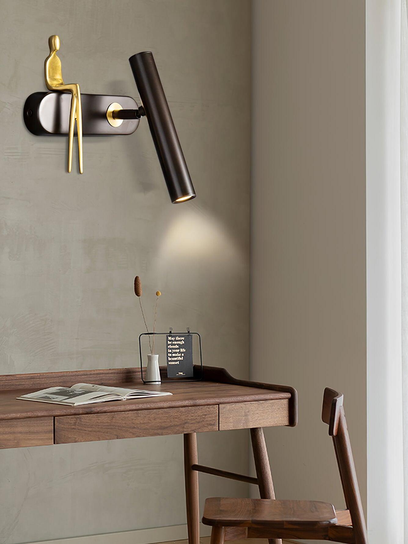 Black wall light with gold seated figure on the left and tilted tube on the right above a wooden desk and open book