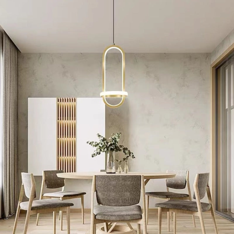 One gold Nyla oval pendant above a round wooden dining table with grey chairs and leafy branches in a vase
