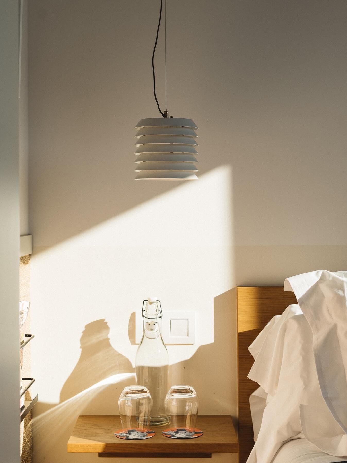 Unlit white layered pendant above bedside shelf and bed linens in sunlight