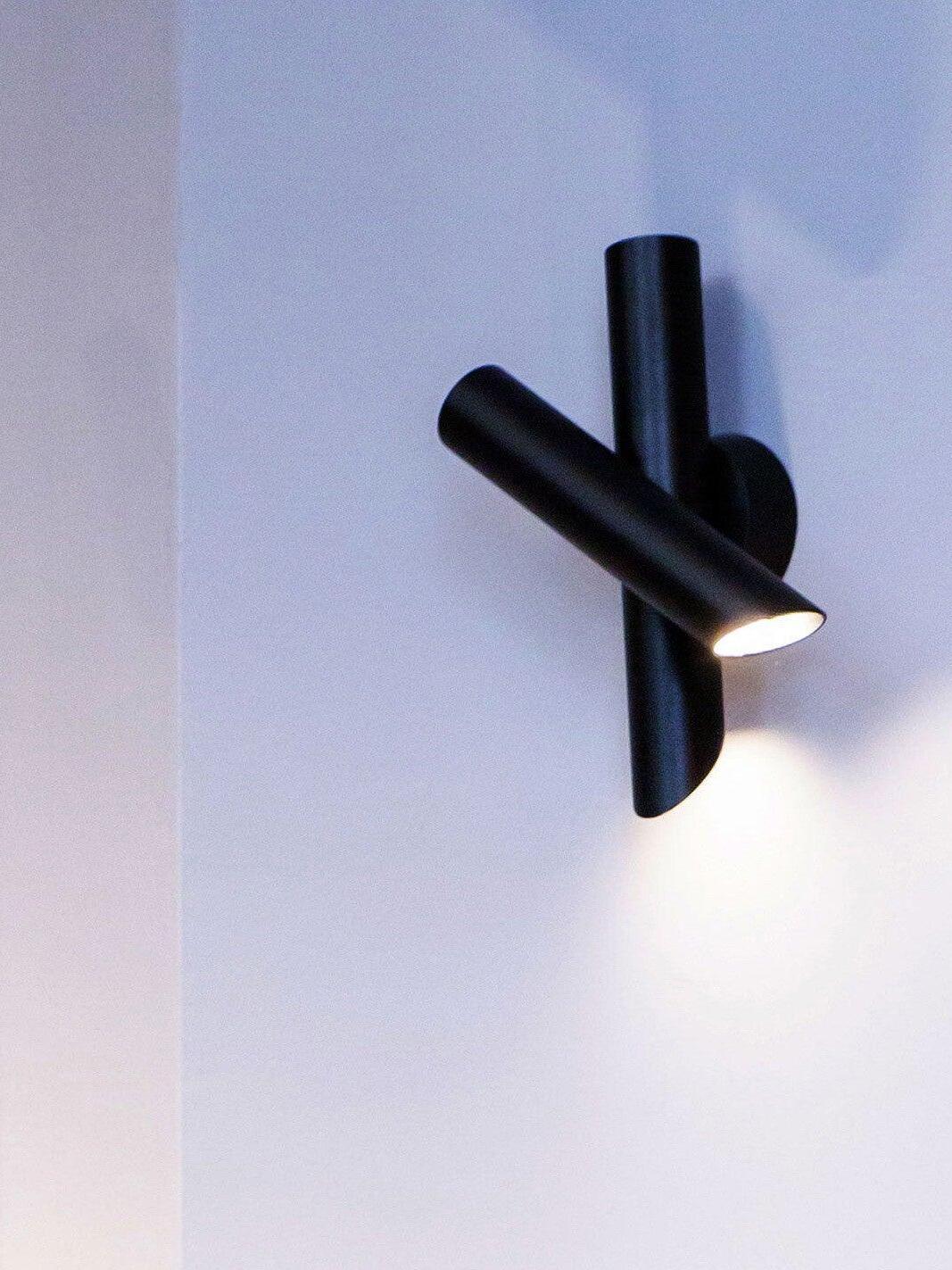 Black crossed-tube wall light glowing on white wall