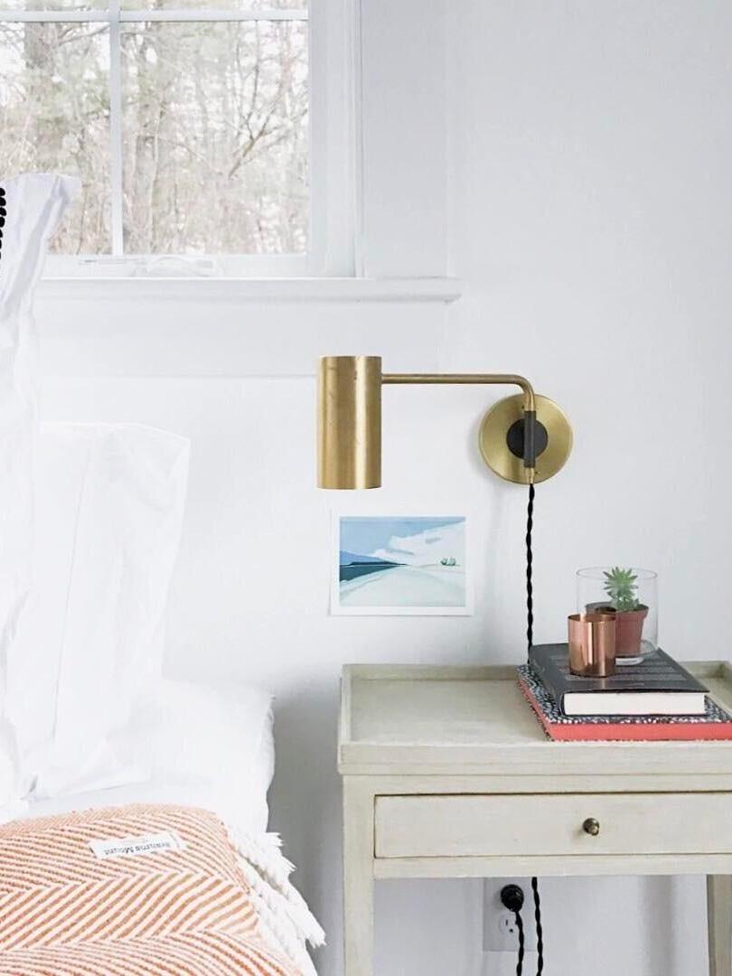 Brass-tone wall light above white bedside cabinet by window