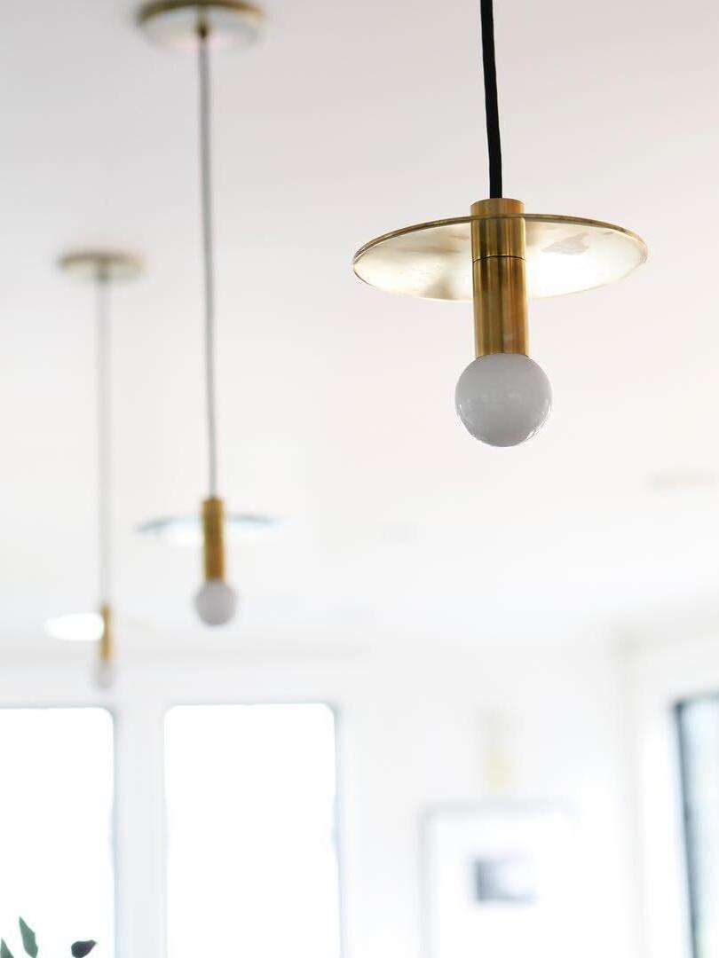 Brass-tone disc pendant with exposed globe bulb in front of two blurred pendants