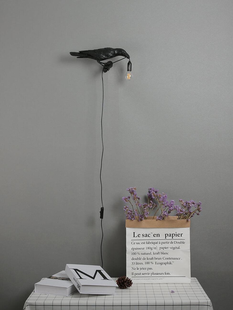 Black Bird wall light with an inline cord switch above books and a bag of flowers on a checked table
