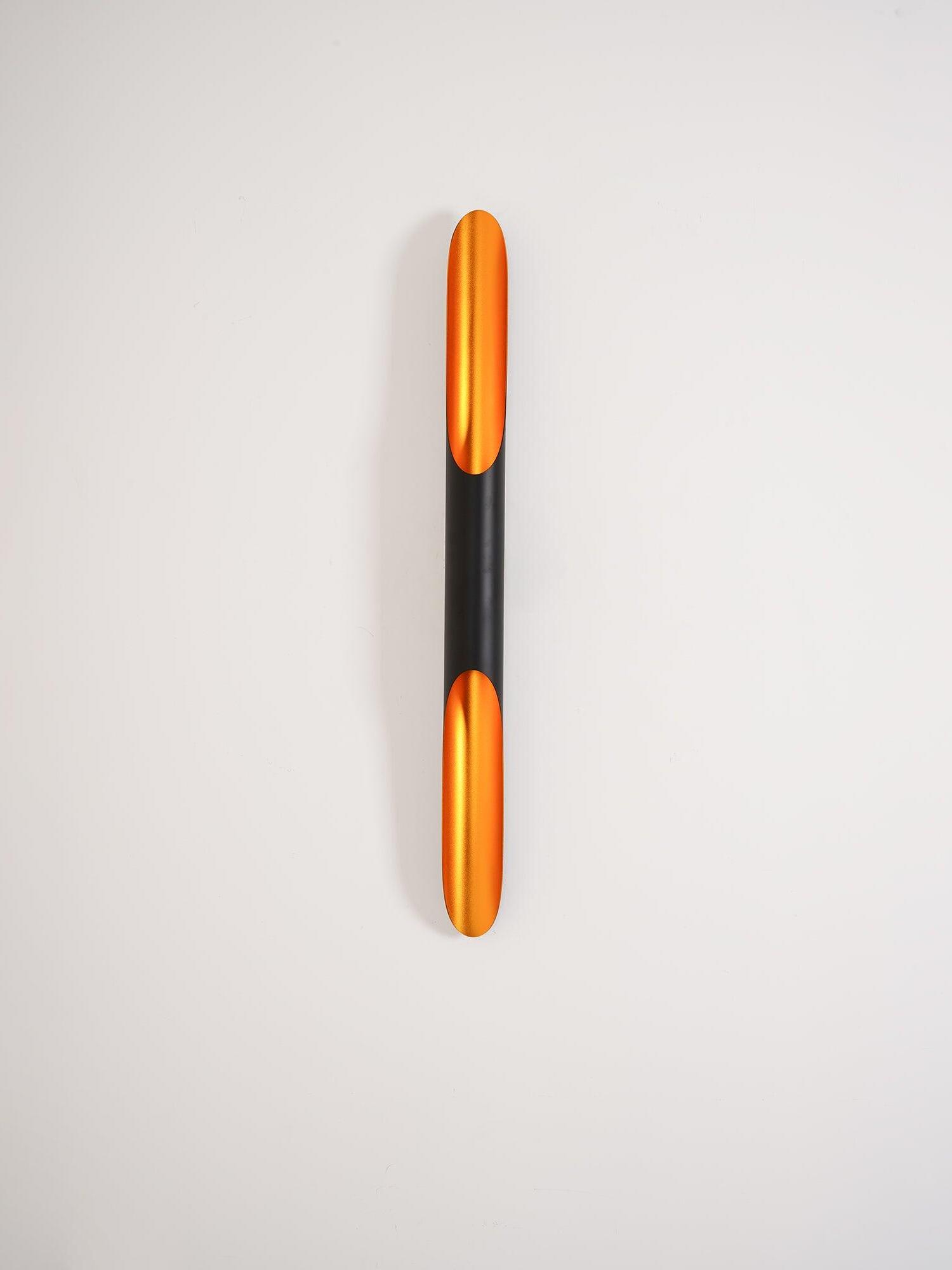 Single black tube wall light with illuminated gold-coloured openings against a pale grey wall