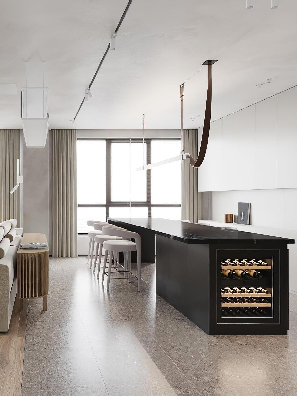 White linear light bar with a brown curved strap end above a black kitchen island with a wine rack and pale chairs