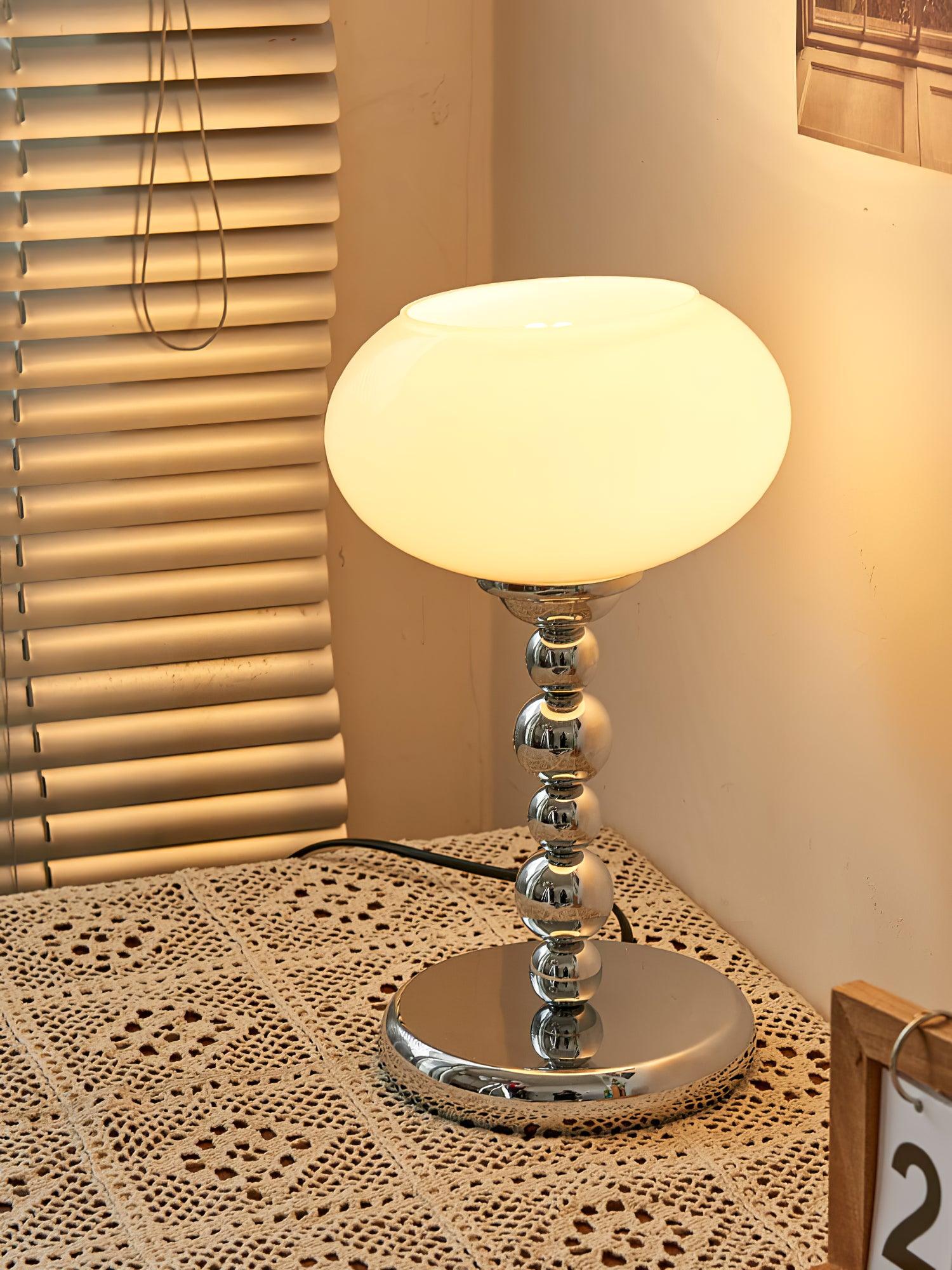 Lit white glass table lamp beside window blinds on lace cloth, with a rounded chrome stem, circular base and black cord