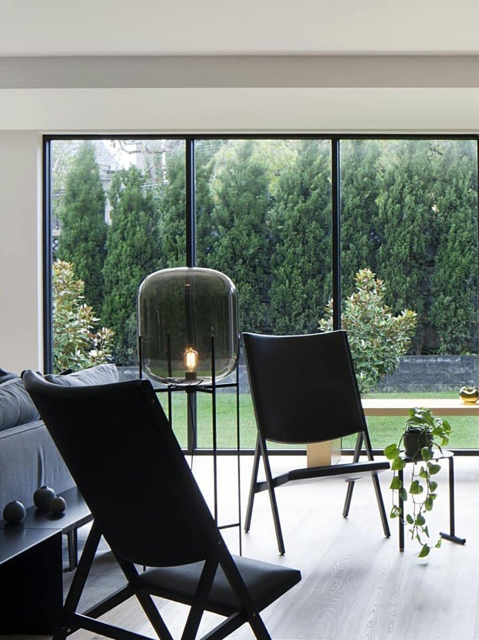 Grey glass lamp beside lounge chairs near a broad garden-facing window.
