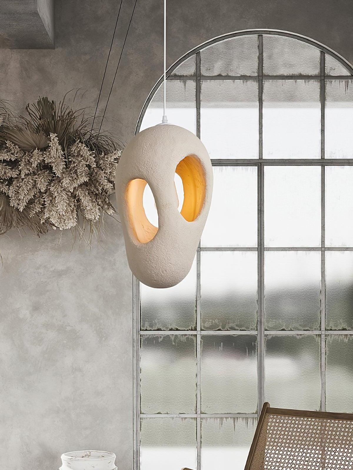 Lit two-opening sculptural pendant beside an arched metal-frame window, dried branches and partly shown woven wood chair