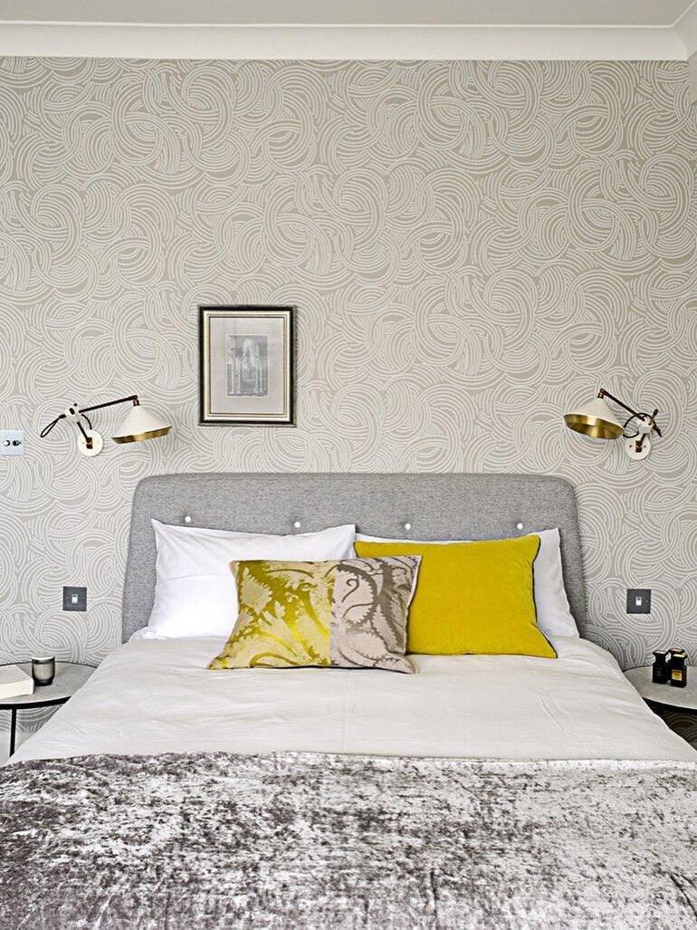 Two white cone wall lights beside a gray headboard with yellow cushions on a patterned wall