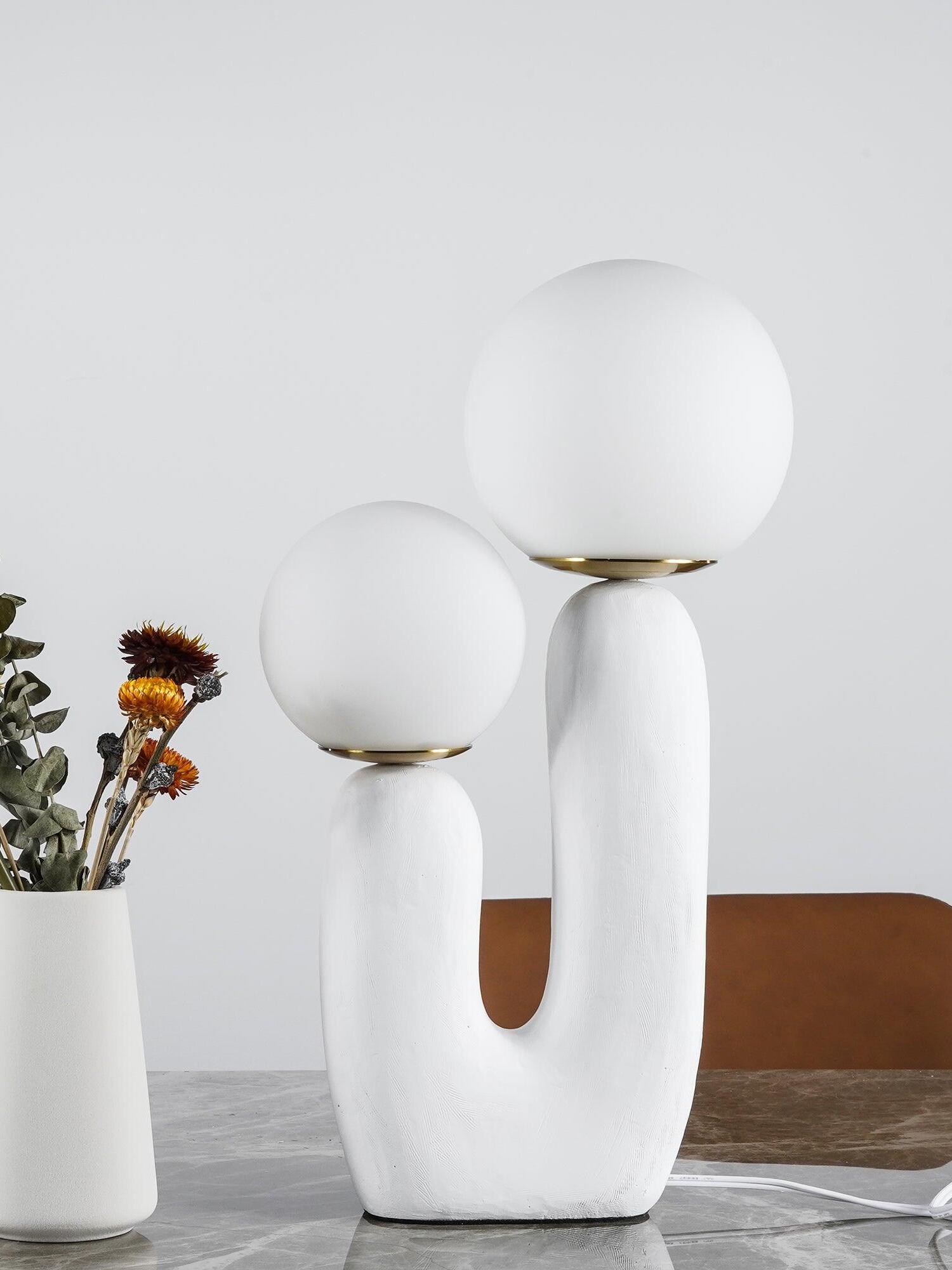 White Oo Rough twin-globe lamp on marble beside a white vase of dried flowers, with brown chair behind