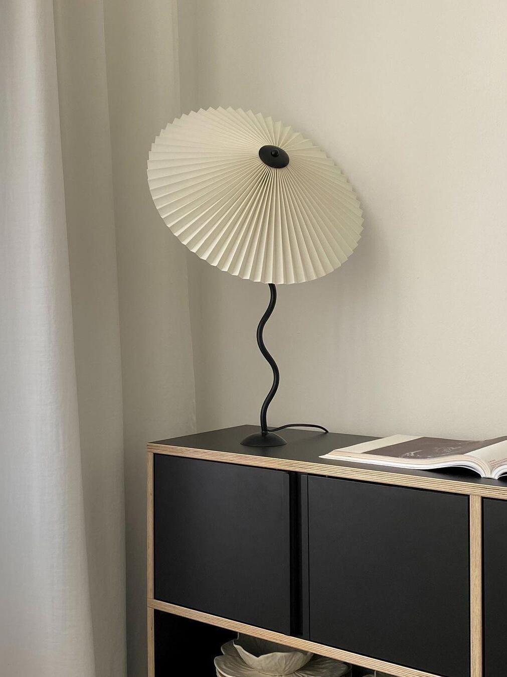 Angled white pleated shade on a black wavy stem above a black-and-wood cabinet beside an open magazine