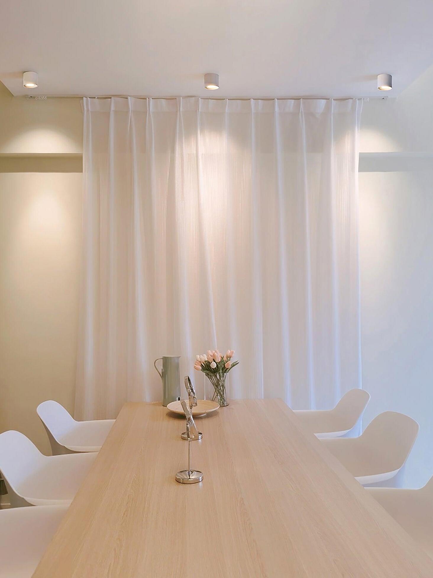 Three white ceiling lights near sheer curtains above a wooden dining table with white chairs, tulips and green pitcher