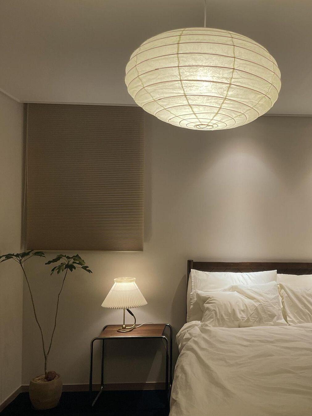 Illuminated broad paper lantern above a bed with white pillows, bedside pleated lamp and a tall plant