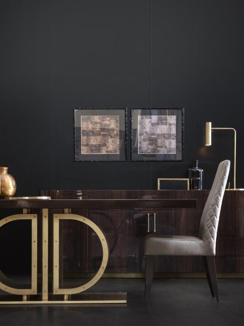 Slim gold-tone lamp on rear desk in dark room with framed art and chair