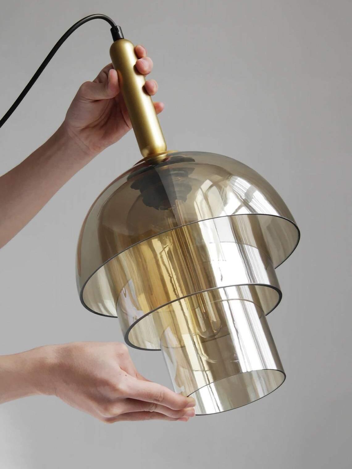 Hands holding a three-tier glass pendant by its gold-tone top fitting and lower glass edge, with black cable above