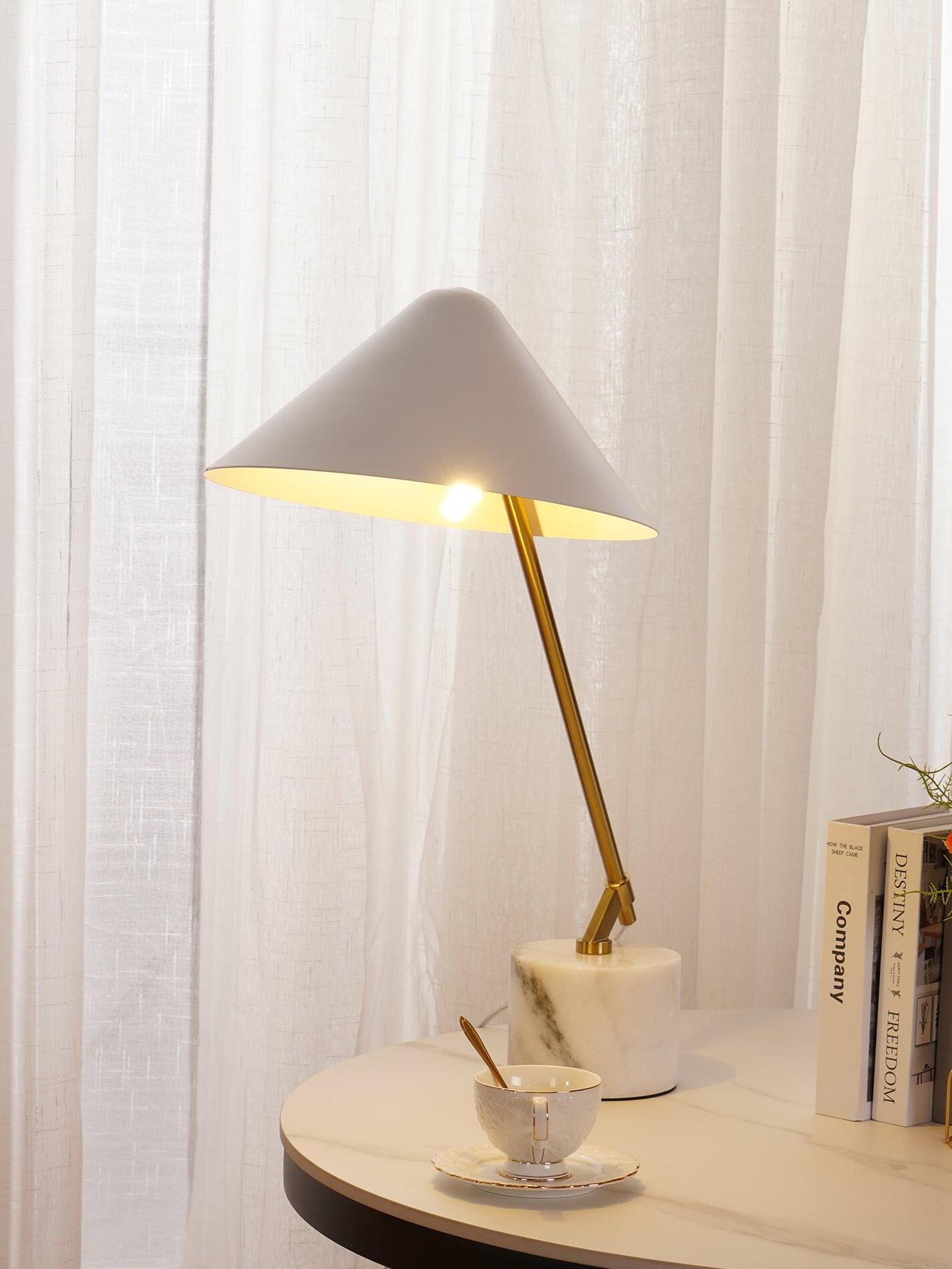 Lit white table lamp with gold-tone arm beside books and a cup near pale curtains