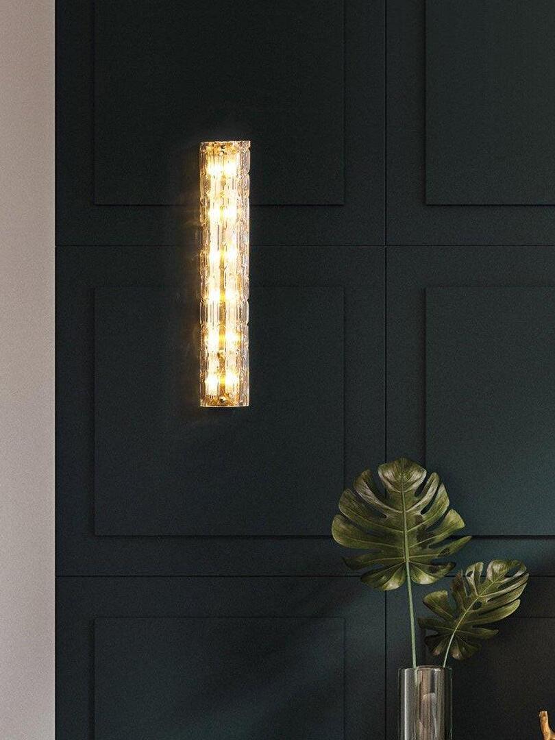Warm-lit glass wall light on dark panels beside greenery