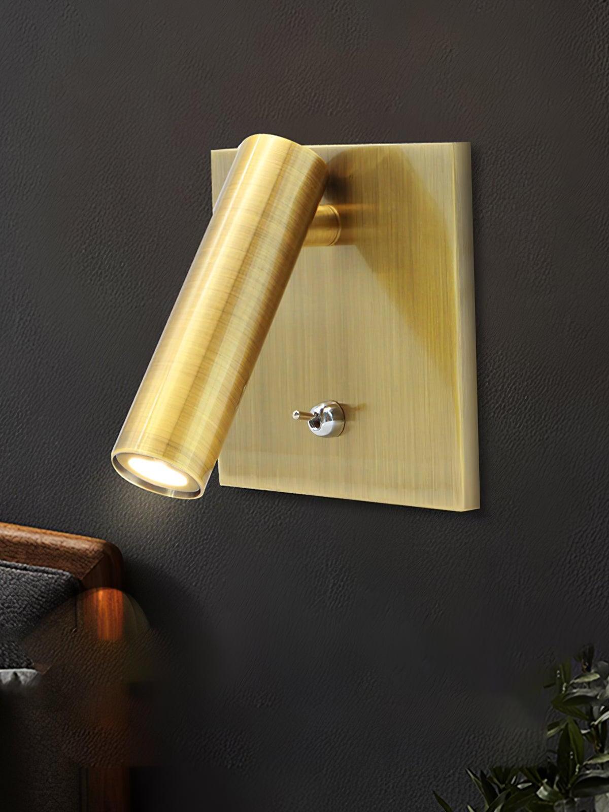 Enna gold-tone lit cylindrical wall light with square plate and silver toggle on dark wall above wood-framed furniture and plant