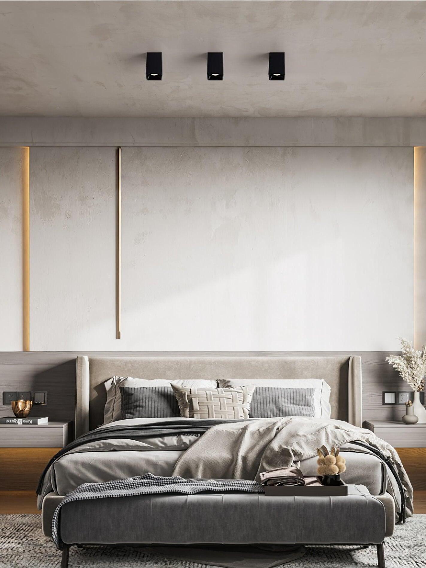 Three black ceiling spots above a gray bed and bench against a pale wall with vertical lighting